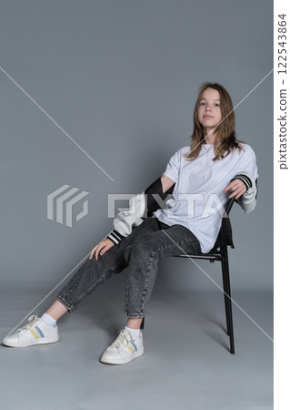 A teenage girl sits casually in a chair, her gaze steady and assured. Her posture reflects the self-confidence and ease of todays youth, embracing a minimalist style. 122543864