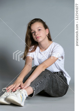 A teenage girl sits thoughtfully on the floor with her hands resting on her legs. This casual portrait highlights the simplicity and authenticity of modern youth culture. A teenage girl sits thoughtfully on the floor with her hands resting on her legs. This casual portrait highlights the simplicity and authenticity of modern youth culture. 122543877