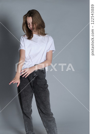 A young girl dressed in a white t-shirt and jeans looks down while standing calmly. This portrait emphasizes a quiet moment of reflection, showcasing natural beauty and minimalism. 122543889