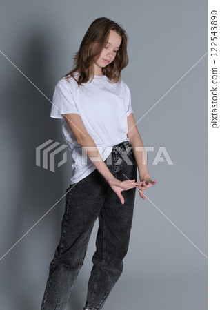 A young girl looks down as she stands with her arms relaxed, wearing a simple white t-shirt and jeans. Her natural posture and casual style convey a sense of calm and introspection. A young girl looks down as she stands with her arms relaxed, wearing a simple white t-shirt and jeans. Her natural posture and casual style convey a sense of calm and introspection. 122543890