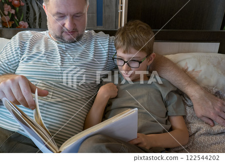 Father hugs his little school-age son and reads funny paper literature. Happy little boy reading a book, enjoying his free weekend with his joyful father on the sofa Father hugs his little school-age son and reads funny paper literature. Happy little boy reading a book, enjoying his free weekend with his joyful father on the sofa 122544202