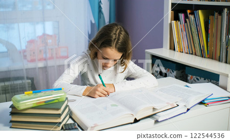Young girl sits at her desk in the evening, deeply engaged in homework, surrounded by study materials 122544536