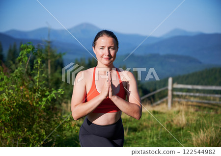 Woman practicing yoga and meditating outdoors in the mountains in serene, natural setting. Portrait of female performing yoga pose, with backdrop of beautiful mountain landscape at sunrise or sunset. 122544982