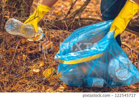A gloved hand picks up a plastic bottle from the ground and places it in a trash bag. A volunteer man removes trash scattered in the forest. Eco, saving nature, eco activism. 122545717