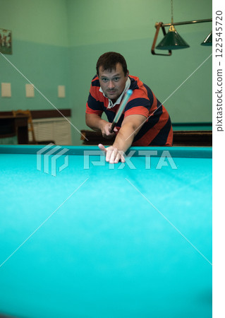Cute man aiming a cue at a billiard ball. In the evening, a man relaxes and relaxes while playing billiards. A man hits the ball with a cue, aiming for a pocket on a billiard table. 122545720