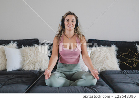 Smiling yoga woman in headphones sitting in lotus position on couch enjoy meditation 122548390