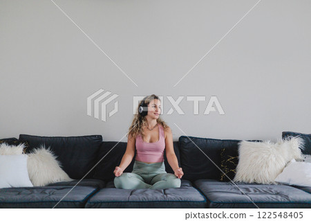 Smiling yoga woman in headphones sitting in lotus position on couch enjoy meditation Smiling yoga woman in headphones sitting in lotus position on couch enjoy meditation 122548405
