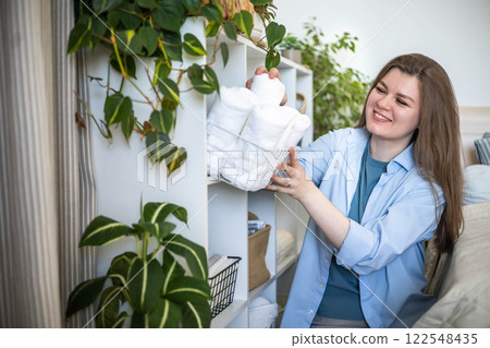 Modern housewife putting neatly folded towels, pillowcases, duvet covers into wicker natural basket Modern housewife putting neatly folded towels, pillowcases, duvet covers into wicker natural basket 122548435