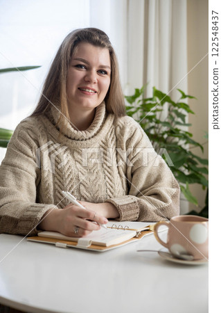 Dreaming plus size woman relaxing at comfortable room with houseplant writing notes in diary 122548437