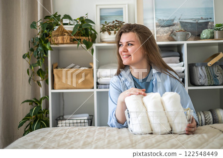 Modern housewife putting neatly folded towels, pillowcases, duvet covers into wicker natural basket Modern housewife putting neatly folded towels, pillowcases, duvet covers into wicker natural basket 122548449