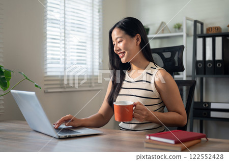 A satisfied Asian businesswoman is enjoying her coffee while working on her laptop in her office. A satisfied Asian businesswoman is enjoying her coffee while working on her laptop in her office. 122552428
