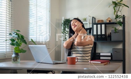 An uncomfortable, tired Asian businesswoman sits at her desk in the office, massaging her neck. An uncomfortable, tired Asian businesswoman sits at her desk in the office, massaging her neck. 122552438