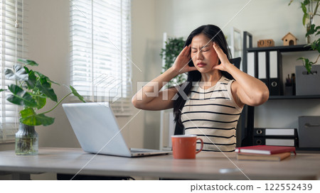 A frustrated, tired Asian businesswoman sits in front of a computer, massaging her temples. 122552439