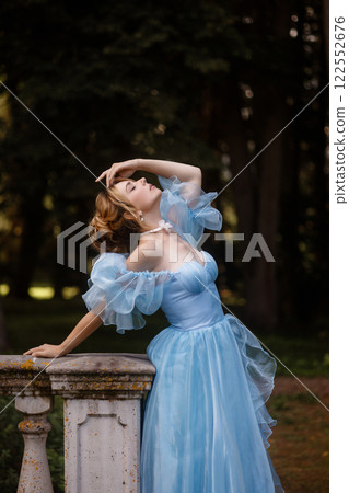 Romantic Woman in a Light Blue Gown Posing on a stone balustrade in a picturesque garden. Romantic Woman in a Light Blue Gown Posing on a stone balustrade in a picturesque garden. 122552676