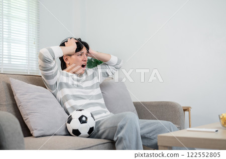A shocked Asian man is watching his favorite football team lose to another team on TV at home. 122552805
