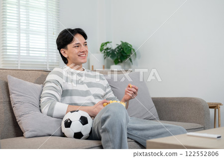 A relaxed Asian man enjoys eating popcorn while watching his favorite football team's match on TV. 122552806