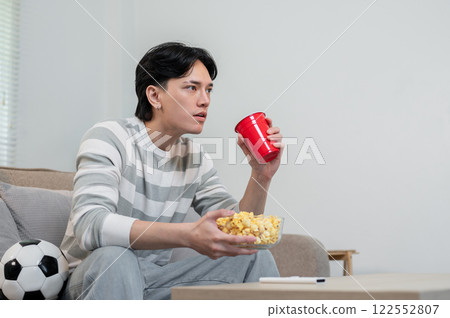 An excited Asian man, focused on his favorite football team's match on TV, sitting on the sofa. 122552807