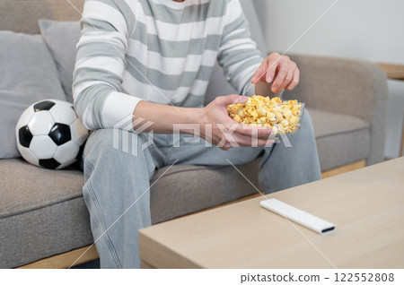 A close-up of a man enjoys eating popcorn while watching his favorite football team's match on TV. 122552808
