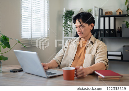 A handsome, smart Asian businessman is working on his laptop at a desk in his minimalist office. 122552819
