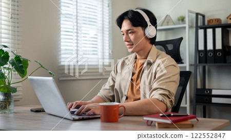 A creative Asian businessman is listening to music on his headphones while working on his laptop. 122552847