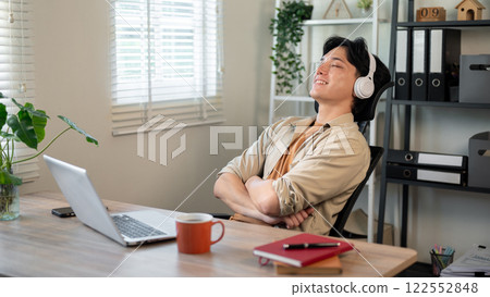A relaxed Asian businessman is leaning back in his chair, enjoying the music on his headphones. A relaxed Asian businessman is leaning back in his chair, enjoying the music on his headphones. 122552848