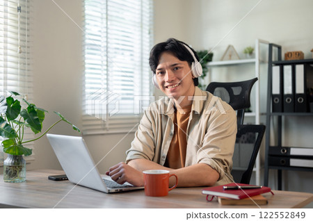 A creative Asian businessman is sitting in front of a laptop in his office, smiling at the camera. 122552849