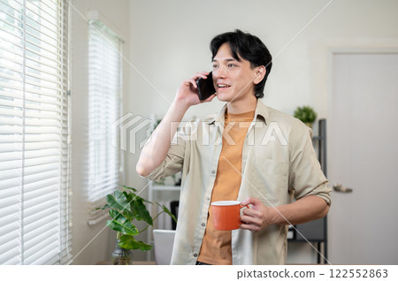 A positive Asian businessman stands in his office with a coffee cup in hand, talking on the phone. A positive Asian businessman stands in his office with a coffee cup in hand, talking on the phone. 122552863