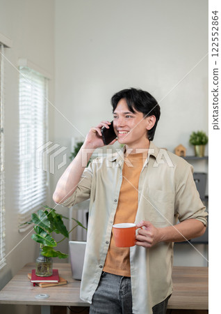 A smiling Asian businessman stands in his office, gazing out the window while talking on the phone. A smiling Asian businessman stands in his office, gazing out the window while talking on the phone. 122552864