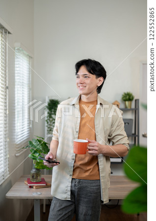 A positive Asian businessman stands in his office with a smartphone and a coffee cup in his hand. A positive Asian businessman stands in his office with a smartphone and a coffee cup in his hand. 122552865