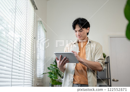 A happy Asian man stands in a room, using a digital tablet, drawing and designing his project. A happy Asian man stands in a room, using a digital tablet, drawing and designing his project. 122552871
