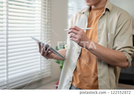 A close-up of an Asian man stands in a room, using a digital tablet, designing his project. 122552872