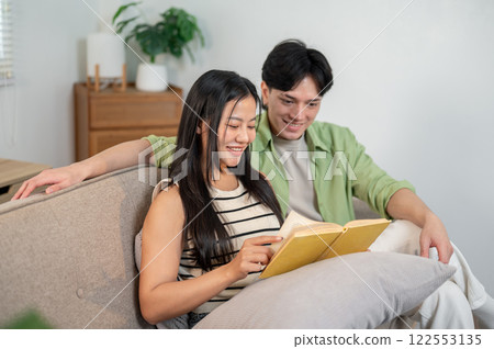 A lovely Asian couple, a boyfriend and girlfriend, enjoy reading a book together on the sofa. 122553135