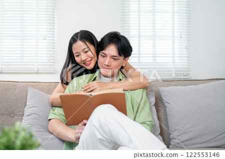 A sweet Asian girl is hugging her boyfriend from behind while he reads a book on the sofa. A sweet Asian girl is hugging her boyfriend from behind while he reads a book on the sofa. 122553146