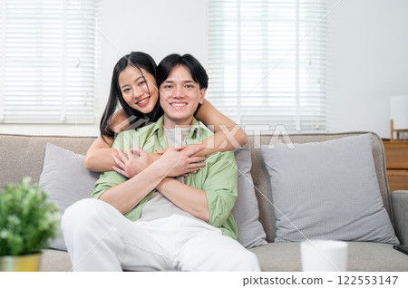 A sweet Asian couple in the living room at home. The girlfriend is hugging her boyfriend from behind A sweet Asian couple in the living room at home. The girlfriend is hugging her boyfriend from behind 122553147