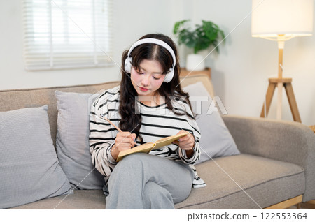 A beautiful Asian woman is deep in contemplation as she writes in her diary on the sofa in her home. 122553364