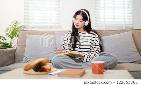 A beautiful young Asian woman in cozy clothes, wearing headphones, writes in her diary on the sofa. A beautiful young Asian woman in cozy clothes, wearing headphones, writes in her diary on the sofa. 122553365