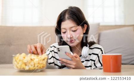 A relaxed Asian woman is enjoying popcorn while reading chats on her smartphone in the living room. 122553410