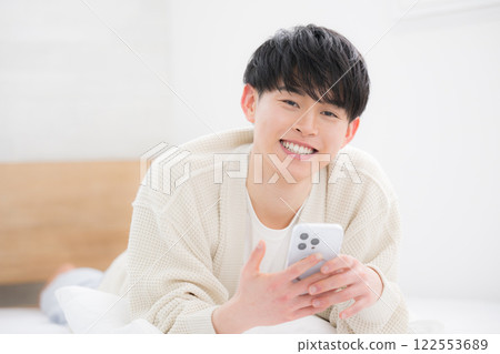 A smiling young man in his 20s lying face down on his bed and using a computer, looking at the camera. Student or freelancer A smiling young man in his 20s lying face down on his bed and using a computer, looking at the camera. Student or freelancer 122553689