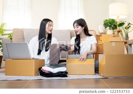 Two Asian female online shop owners are working in the living room, discussing while packing goodies Two Asian female online shop owners are working in the living room, discussing while packing goodies 122553733
