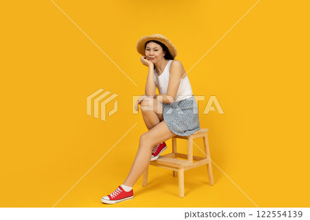 An Asian girl in a summer outfit is sitting on a chair, on a yellow background. An Asian girl in a summer outfit is sitting on a chair, on a yellow background. 122554139