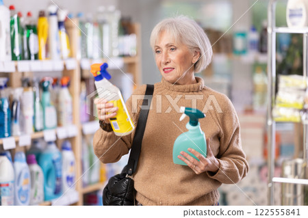 Elderly female shopper carefully selects a spray glass cleaner for windows in home Elderly female shopper carefully selects a spray glass cleaner for windows in home 122555841