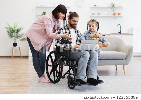 Caucasian family in modern kitchen, mother, father in wheelchair with disability, young daughter with teddy bear. Warm family moment showing love and support. Caucasian family in modern kitchen, mother, father in wheelchair with disability, young daughter with teddy bear. Warm family moment showing love and support. 122555869
