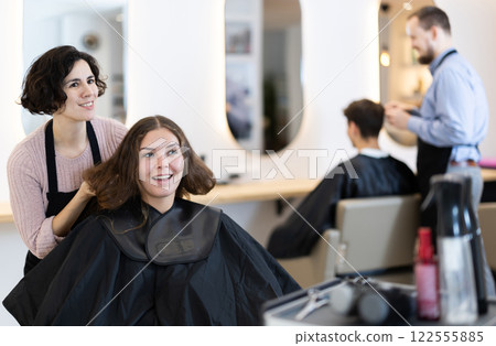 Young woman barbershop employee stands next to female client, discussing details of haircut Young woman barbershop employee stands next to female client, discussing details of haircut 122555885