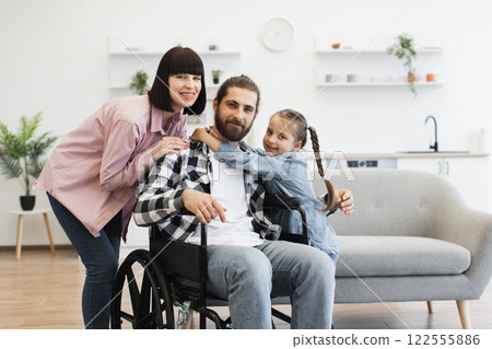 Portrait of Caucasian family with father in wheelchair, mother, and young daughter in kitchen. Warm and supportive family bond captured in comfortable, modern home environment. 122555886