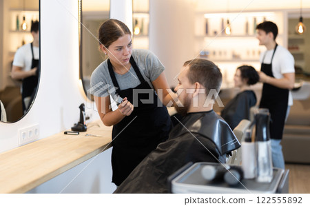 Young woman barbershop employee stands next to male client, discussing details of haircut 122555892