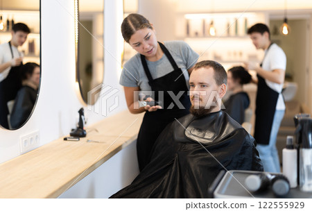 Young woman barbershop employee stands next to male client, discussing details of haircut 122555929