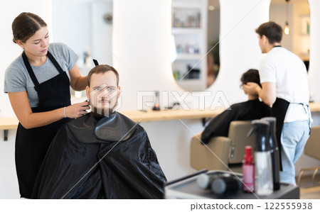 Young woman barbershop employee stands next to male client, discussing details of haircut 122555938