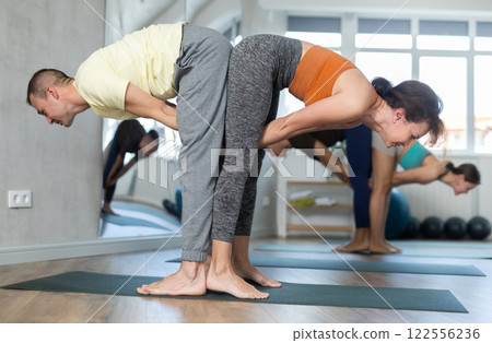 Women and men performing paired yoga exercises during group training at gym. Fitness and pair Hatha yoga concept. Women and men performing paired yoga exercises during group training at gym. Fitness and pair Hatha yoga concept. 122556236