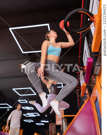 Young woman throwing ball in basket high jumping in trampoline arena Young woman throwing ball in basket high jumping in trampoline arena 122556423