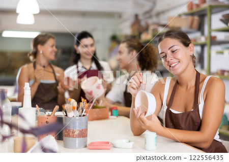 Young woman sits near table and decorates clay ceramic craft vase, plate Young woman sits near table and decorates clay ceramic craft vase, plate 122556435